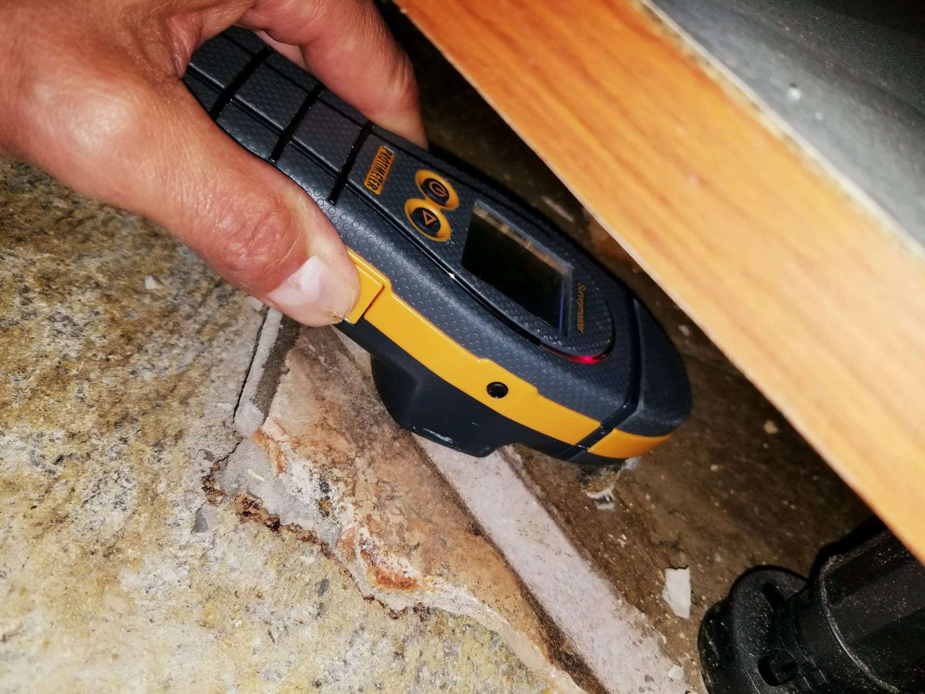 Hand holding a pin moisture meter against a baseboard beside a cabinet